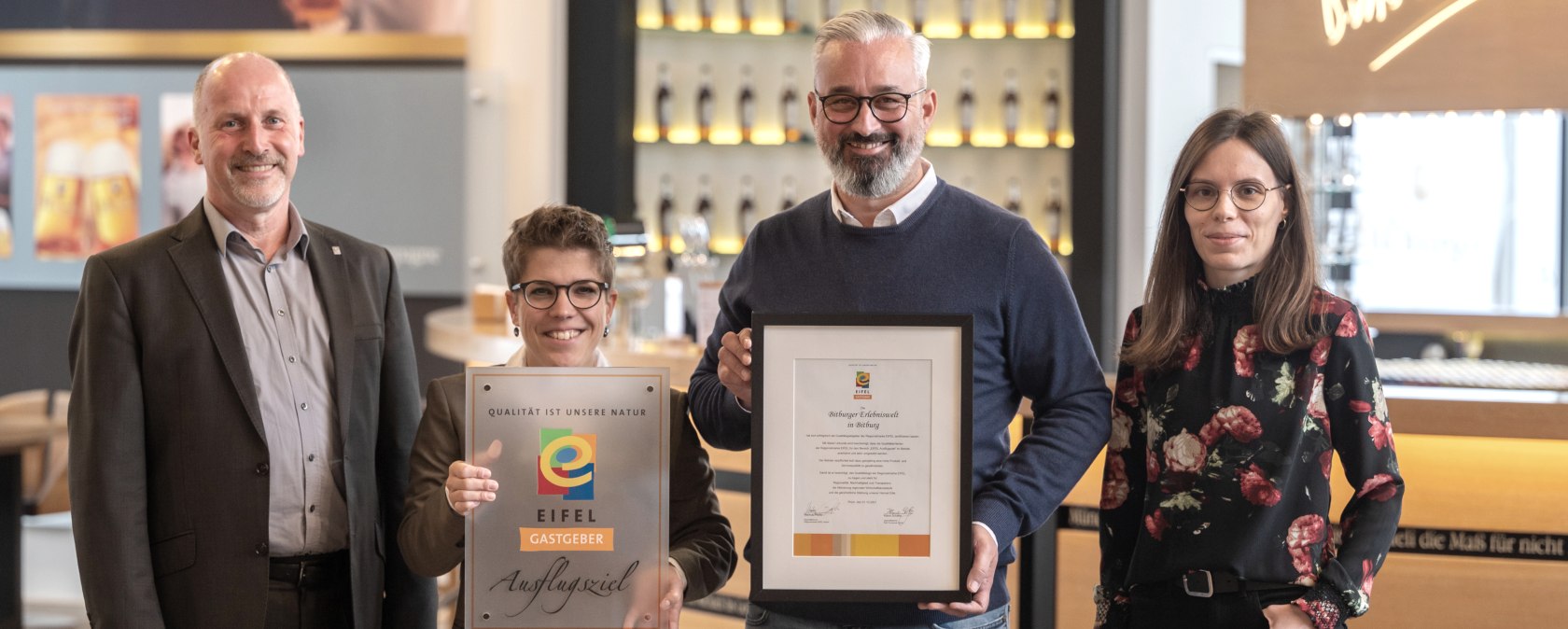 Marco Boor, Leiter Regionales Marketing und Sponsoring Bitburger, und Susanna Schneider, Gästebegleiterin und Q-Coach der Bitburger Erlebniswelt, nehmen die EIFEL Gastgeber-Plakette von Markus Pfeifer und Alina Wonner in Empfang, © Bitburger / Harald Tittel