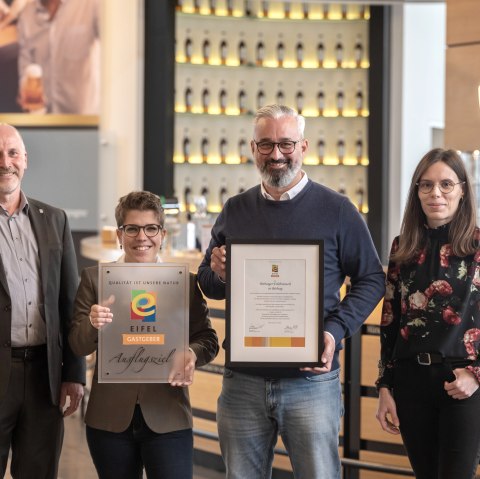 Marco Boor, Leiter Regionales Marketing und Sponsoring Bitburger, und Susanna Schneider, Gästebegleiterin und Q-Coach der Bitburger Erlebniswelt, nehmen die EIFEL Gastgeber-Plakette von Markus Pfeifer und Alina Wonner in Empfang, © Bitburger / Harald Tittel